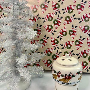 Puede incluir: Un calentador de aceite de cerámica blanca con un diseño pintado de gansos con cintas rojas y coronas verdes, acompañado de un pequeño árbol de Navidad artificial de oropel plateado y papel de regalo con un patrón de Papá Noel.