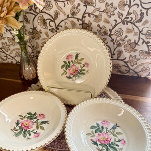 Pode incluir: Um conjunto de três tigelas de cerâmica creme com um desenho floral de rosas no centro. As tigelas têm uma borda dourada e uma borda ondulada. As tigelas são exibidas em uma superfície decorativa.