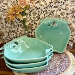 May include: A set of four light green ceramic dishes shaped like apples, each featuring a molded leaf detail at the top. The dishes have a smooth, glossy finish and are stacked together, with one additional matching dish standing upright behind them.
