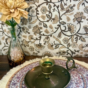 Peut inclure: Un bougeoir en laiton vintage avec une base ronde et une poignée. Le bougeoir est posé sur un tapis rond décoratif à motifs floraux. Un vase avec des fleurs est à l'arrière-plan.