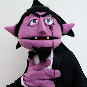 May include: A purple puppet with a black beard, wearing a black tuxedo, white shirt, and a monocle. The puppet has large ears, a prominent nose, and two visible fangs. The puppet is holding its hand up, and has a black bow tie.