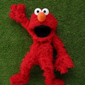 May include: A plush red character with large white eyes, an orange nose, and a wide black mouth. The character is lying on a green surface, with one arm raised in a waving gesture. The toy is a popular children's character.