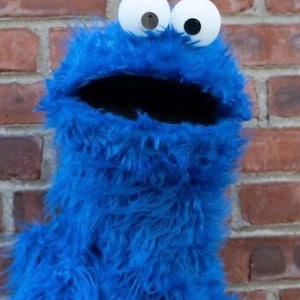 Könnte beinhalten: Eine blaue, pelzige Puppe mit großen weißen Augen und offenem Mund. Die Puppe hat eine zottelige Textur und steht vor einer Backsteinmauer. Die Puppe ist eine bekannte Figur aus einer Kinderserie.