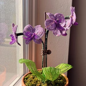 May include: A beaded orchid plant featuring light purple and dark violet petals, green beaded leaves, and a central stem supported by a metal clip. The plant is potted in a terracotta-coloured container filled with green moss, creating a detailed floral sculpture.
