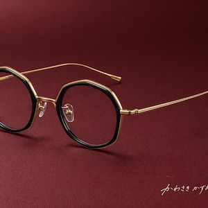 May include: Spectacles featuring a gold-tone metal frame with black rims around the hexagonal lenses. The thin temples are gold-tone metal, and the bridge includes adjustable nose pads. The Japanese text "かわさきかずお" is visible in the corner.
