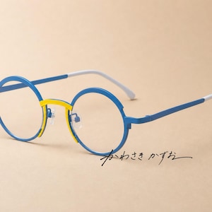 Puede incluir: Gafas redondas con montura azul brillante, puente amarillo y puntas de patillas blancas. Los cristales transparentes son circulares y la montura incluye el texto japonés "かわさき かずお" escrito en negro en la esquina inferior derecha.