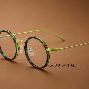 May include: Round spectacles featuring a lime green metal frame and thin temples. The circular rims display a multi-coloured pattern of teal, brown, and orange stripes. Clear nose pads are attached to the bridge. The text "かわさき かずお" is visible.