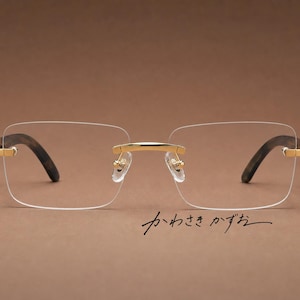 Puede incluir: Gafas sin montura con lentes transparentes rectangulares, un puente de metal dorado y patillas con patrón marrón oscuro. La montura incluye bisagras doradas y almohadillas nasales transparentes. El texto japonés "かわさき かずお" es visible.