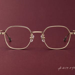 Puede incluir: Gafas de metal en tono dorado con un diseño de montura hexagonal fina, puente, almohadillas nasales ajustables y patillas delgadas. La montura incluye detalles texturizados en las bisagras y el texto japonés "かわさきかずお" en la esquina.