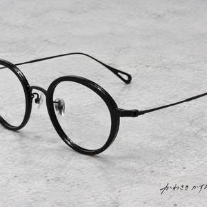 Puede incluir: Gafas negras de montura redonda con un marco circular grueso, patillas de metal finas y lentes transparentes. Las gafas incluyen almohadillas nasales ajustables y un detalle de bucle en el extremo de cada patilla, con texto japonés visible.