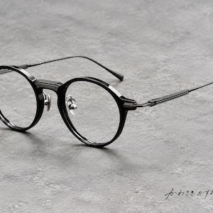 Puede incluir: Gafas negras de montura redonda con borde grueso de acetato, puente metálico y varillas finas y texturizadas. Las gafas incluyen almohadillas nasales ajustables y lentes transparentes. El texto japonés "かわさきかずお" es visible en la esquina inferior derecha.