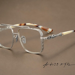 Puede incluir: Gafas de metal plateado con diseño de doble puente, monturas cuadradas con detalles grabados intrincados y varillas que terminan en un patrón de carey de acetato. El texto "かわさき かずお" es visible en la imagen.