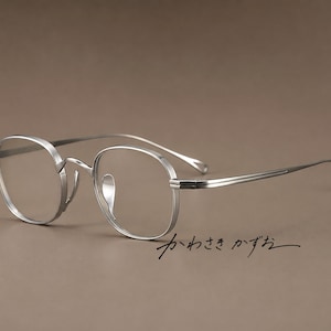 Puede incluir: Gafas de metal plateado con monturas redondeadas y patillas finas y delgadas. Las gafas cuentan con lentes transparentes y almohadillas nasales ajustables. El texto japonés "かわさき かずお" está escrito en negro bajo la montura.