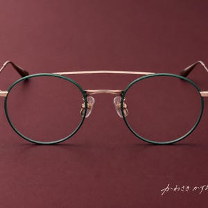May include: Round spectacles featuring a thin rose gold metal frame with dark green rims and a double bridge design. The temples have a textured finish, and the frame includes clear nose pads. The text "かわさきかずお" is visible in the corner.