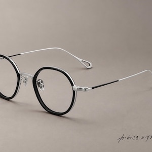 Può includere: Occhiali rotondi con bordi neri e una montatura, ponte e aste sottili in metallo argentato. Le estremità delle aste presentano un design ritagliato distintivo. Il testo giapponese "かわさきかずお" è visibile nell'angolo in basso a destra.