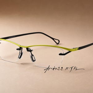 Puede incluir: Gafas semi-al aire con una barra superior de color verde lima brillante y patillas negras finas. Las lentes son transparentes y no tienen montura en la parte inferior. El texto japonés "かわさき かずお" aparece en letra negra abajo a la derecha.