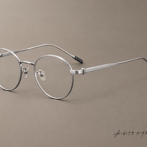 May include: Silver-toned round spectacles featuring thin metal frames, a bridge with adjustable nose pads, and temples with a textured, ribbed pattern. The temple tips are finished with black accents. The Japanese text "かわさきかずお" is visible.