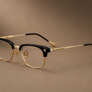 Könnte beinhalten: Brille mit schwarzem Browline-Rahmen sowie goldfarbenen Metallrändern und Bügeln. Der Rahmen ist an den vorderen Ecken mit kleinen silberfarbenen Fleur-de-Lis-Akzenten versehen und weist dekorative Gravuren entlang der dünnen goldfarbenen Bügel auf.