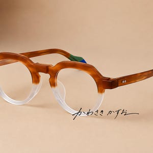 May include: Round spectacles with a two-tone frame featuring a matte brown upper section and a clear lower section. The temples have green and blue tips. The Japanese text "かわさき かずお" is printed in black near the bottom right lens.