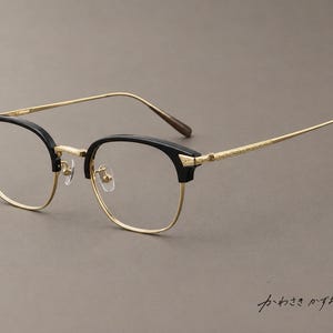 Op de afbeelding: Bril met een zwart browline montuur, goudkleurige metalen randen en getextureerde goudkleurige pootjes. De brug en neuspads zijn van goudkleurig metaal. De Japanse tekst "かわさきかずお" is rechtsonder in de afbeelding zichtbaar.