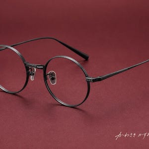 以下が含まれることがあります： リムとテンプルに質感のある仕上げが施された、マットブラックのメタルフレームの丸眼鏡。透明なレンズ、調整可能なノーズパッド、細いテンプルが特徴です。隅には日本語の「かわさきかずお」という文字が見えます。