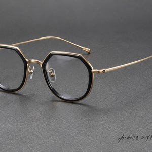 Könnte beinhalten: Brille mit einem goldfarbenen Metallrahmen und schwarzen Rändern um die achteckigen Gläser. Die dünnen, zierlichen Bügel erstrecken sich zu den Seiten und der Rahmen verfügt über verstellbare Nasenpads. Der japanische Text "かわさきかずお" ist sichtbar.