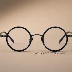 May include: Black round-frame eyeglasses featuring thin metal rims and temples with a textured pattern. The frames have a bridge connecting the circular lenses, and the Japanese text "かわさきかずお" is written in black script below the right lens.
