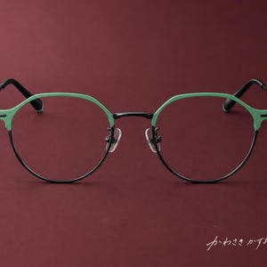 Può includere: Occhiali dal design bicolore con bordi superiori verde menta e bordi inferiori neri sottili. La montatura include un ponte nero, naselli regolabili e aste nere. Il testo giapponese "かわさきかずな" è visibile nell'angolo in basso a destra.