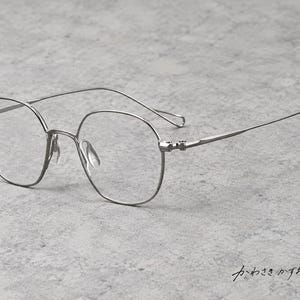 Può includere: Occhiali in metallo color argento con montature sottili e arrotondate e aste slanciate. Gli occhiali hanno lenti trasparenti e naselli regolabili. Il testo giapponese "かわさきかずお" è visibile nell'angolo in basso a destra.
