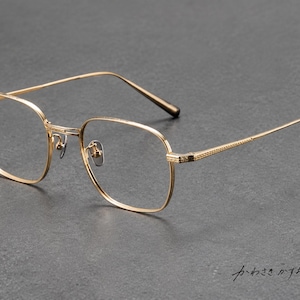 Può includere: Occhiali in metallo color oro con montatura di forma quadrata e bordi arrotondati. Le aste e il ponte presentano un motivo a tratteggio incrociato finemente testurizzato. La montatura include naselli regolabili e aste sottili con punte scure.