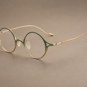 Puede incluir: Gafas redondas con una montura fina de metal dorado y un degradado verde oscuro en los bordes superiores. Las gafas incluyen lentes transparentes, almohadillas nasales ajustables y varillas delgadas y ligeras con una ligera curva al final.