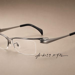 Op de afbeelding: Gunmetal grijze bril met halve rand, een slank metalen montuur, verstelbare neuspads en zwarte uiteinden bij de veren. Het montuur heeft een opvallend scharnierontwerp en de Japanse tekst "かわさき かずお" staat in zwarte letters eronder.