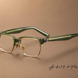 Puede incluir: Gafas de media montura con barras frontales y patillas de acetato verde oliva translúcido, con aros y plaquetas nasales de metal dorado. Las patillas muestran un patrón lineal texturizado. El texto japonés "かわさき かずお" es visible en la esquina inferior derecha.