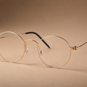 May include: Round spectacles featuring a thin, polished gold-tone metal frame. The design includes clear lenses, a delicate bridge, and slender temples that terminate in black plastic tips. The hinges exhibit a visible coiled metal detail.