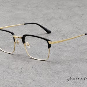 Könnte beinhalten: Brille mit schwarzem Browline-Rahmen, goldfarbenen Metallrändern und dünnen goldenen Bügeln. Der Steg und die Scharniere haben eine strukturierte Oberfläche. Der japanische Text "かわさきかずお" ist unten rechts zu sehen.