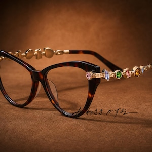 Puede incluir: Gafas de ojo de gato con montura de carey marrón oscuro y patillas de metal dorado. Las patillas están adornadas con una serie de pequeños cristales facetados multicolores en tonos rosa, azul, verde y ámbar. Texto japonés visible.