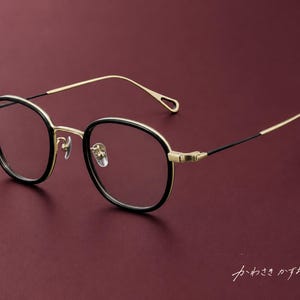 Puede incluir: Gafas con monturas redondas negras y una estructura, puente y patillas finas de metal en tono dorado. Los extremos de las patillas tienen forma de bucle y el texto japonés "かわさきかずお" es visible en la esquina inferior derecha.