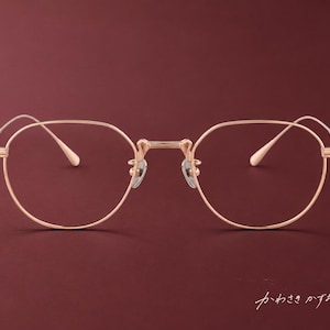 Può includere: Occhiali in metallo color oro rosa con una montatura sottile e arrotondata e naselli regolabili. Le aste presentano un motivo a rilievo. Il testo giapponese "かわさきかずお" è visibile nell'angolo in basso a destra della montatura.