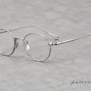Puede incluir: Gafas de metal en tono plateado con monturas redondas, patillas finas y almohadillas nasales integradas. En la esquina inferior derecha aparece el texto japonés "かわさきかずお" en cursiva, identificando al diseñador.