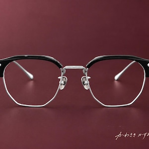 Könnte beinhalten: Browline-Brille mit schwarzem Oberrahmen und silbernem Unterrand. Die Bügel sind silberfarben mit metallischem Finish. Der Steg und die Nasenpads sind silber. Der japanische Text "かわさきかずお" ist in der unteren rechten Ecke zu sehen.