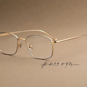 Puede incluir: Gafas de metal en tono dorado con una montura rectangular fina y detalles de borde negro. Las varillas son delgadas y curvadas. El texto japonés "かわさき かずお" está impreso en escritura negra debajo de las gafas en el lado derecho.