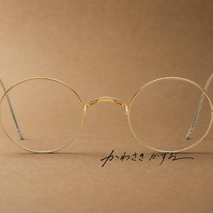 Peut inclure: Lunettes en métal doré dotées de montures circulaires fines et d'un pont délicat. Les branches sont minces avec des embouts texturés. Le texte japonais "かわさきかずお" est imprimé en noir sous le verre droit, indiquant le nom du designer.