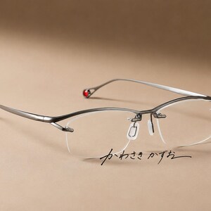 Puede incluir: Gafas semi-al aire de color gris plomo con un diseño de montura fina y elegante y lentes transparentes. Los extremos de las varillas están decorados con pequeñas cuentas rojas redondas. El texto japonés "かわさき かずお" aparece en negro bajo el cristal derecho.