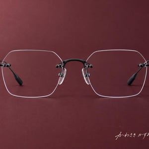 Puede incluir: Gafas sin montura con puente de metal negro y patillas negras finas y texturizadas. Las lentes tienen forma hexagonal. El texto japonés "かわさきかずお" es visible en la esquina inferior derecha del marco.