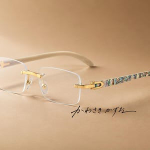 Puede incluir: Gafas sin montura con lentes transparentes rectangulares, puente y bisagras de metal dorado, y patillas con un patrón iridiscente multicolor similar a una concha. El texto japonés "かわさき かずお" es visible bajo la montura.