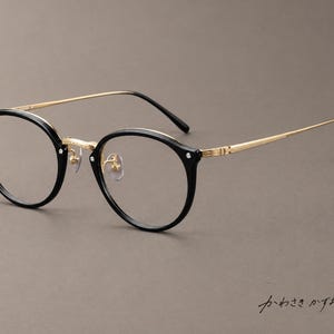 Puede incluir: Gafas redondas con frente de montura de acetato negro, puente de metal dorado y varillas finas de metal dorado texturizado. Las gafas incluyen lentes transparentes, almohadillas nasales ajustables y el texto japonés "かわさきかずお" en la esquina.