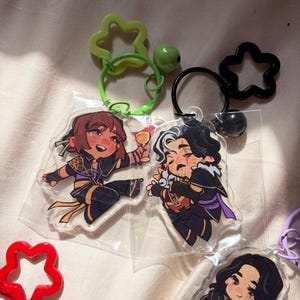 May include: Three acrylic keychains featuring anime-style characters in black and purple outfits. One character holds a golden goblet, another hugs a small creature, and the third plays a guitar. Each keychain has a star-shaped clasp and a small bell.