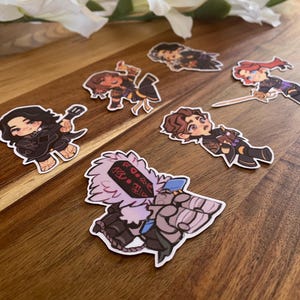 May include: A collection of colorful character stickers on a wooden surface. The stickers feature various cartoon figures in action poses, with detailed outfits and expressions. The stickers are outlined in white, creating a distinct contrast against the wooden background.