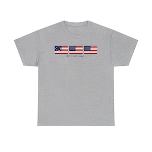 May include: A heather grey short-sleeve t-shirt featuring a graphic of three historical United States flags arranged horizontally. Below the flags, the text 1777 | 1912 | 1960 is printed in a dark font, representing the evolution of the national flag.