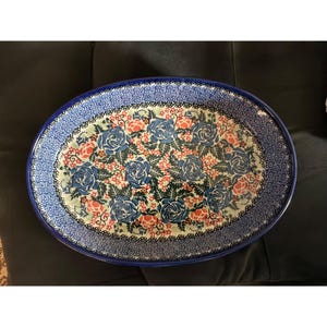 May include: An oval ceramic serving dish featuring a decorative pattern of blue roses and small orange flowers set against a cream background. The rim is adorned with a detailed blue and white geometric border, creating a distinct contrast.